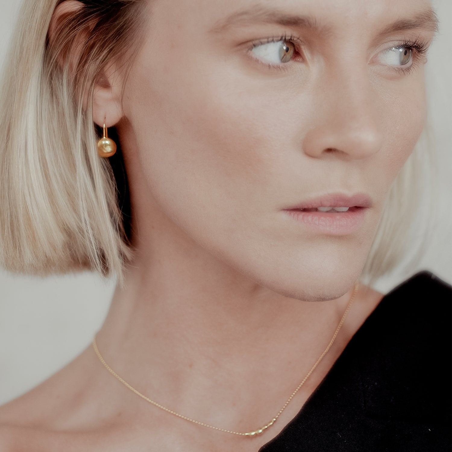 Close-up of a woman wearing gold orb earrings and a mini orb necklace against a neutral background