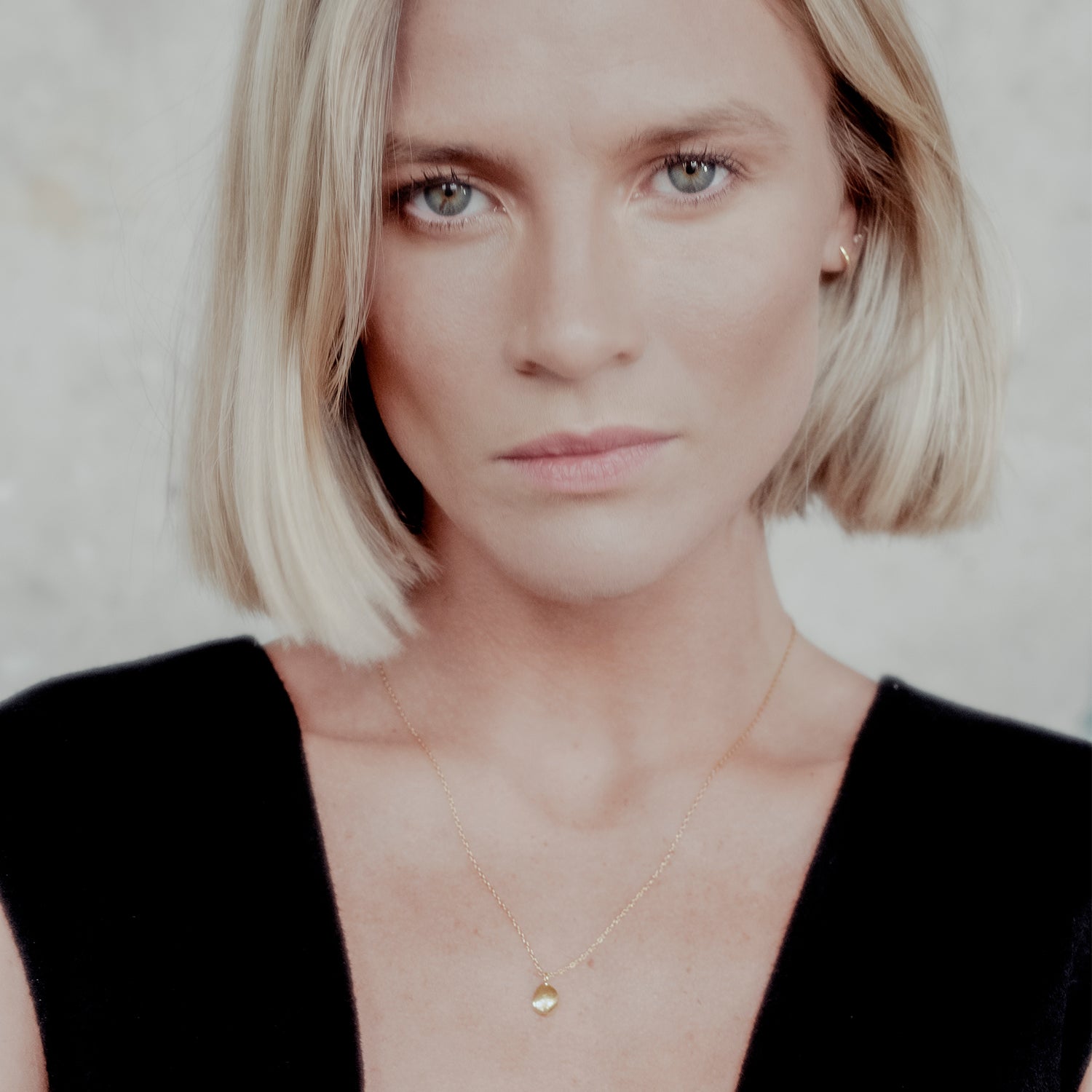 Woman with blonde hair wearing a black top and gold necklace against a neutral background