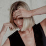 Woman with short blonde hair adjusting her hair against a neutral background, wearing gold paperclip chain necklace and gold birthstone bracelets.
