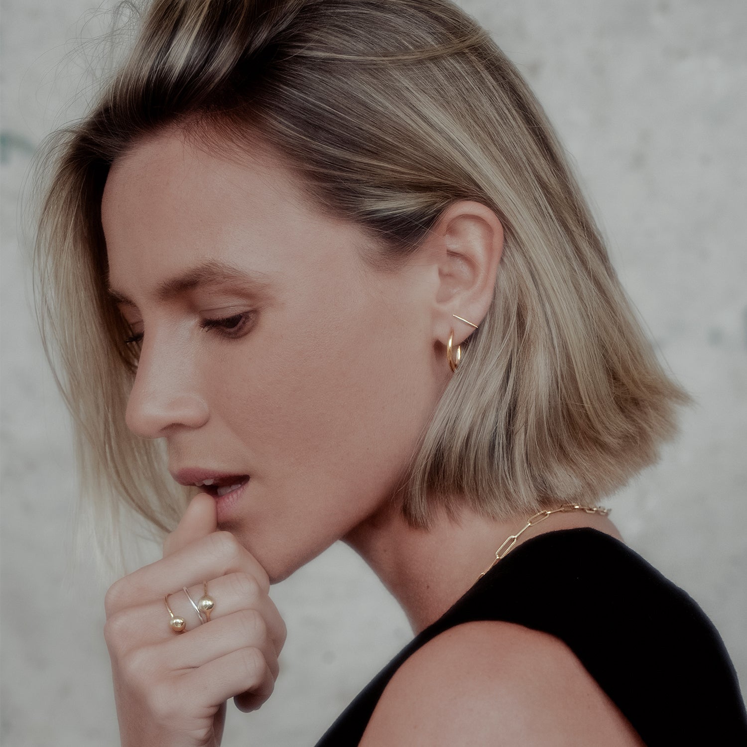 Woman with short blonde hair wearing gold earrings and rings, against a neutral background