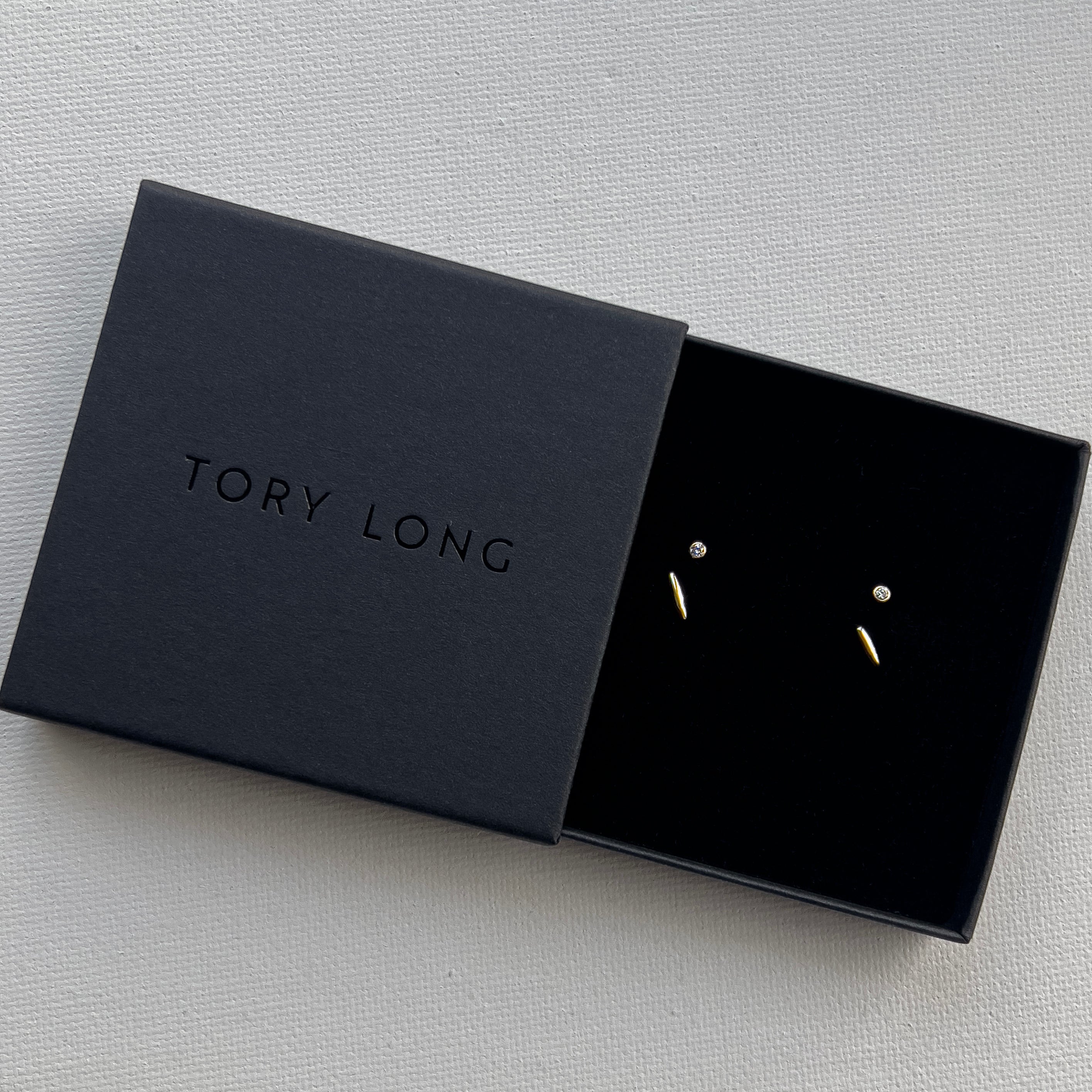 Black jewelry box with 'Tory Long' branding containing a pair of earrings in 9ct gold and salt and pepper diamonds on a light gray background.