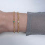 Arm wearing two gold ball chain bracelets set with emerald and diamonds.