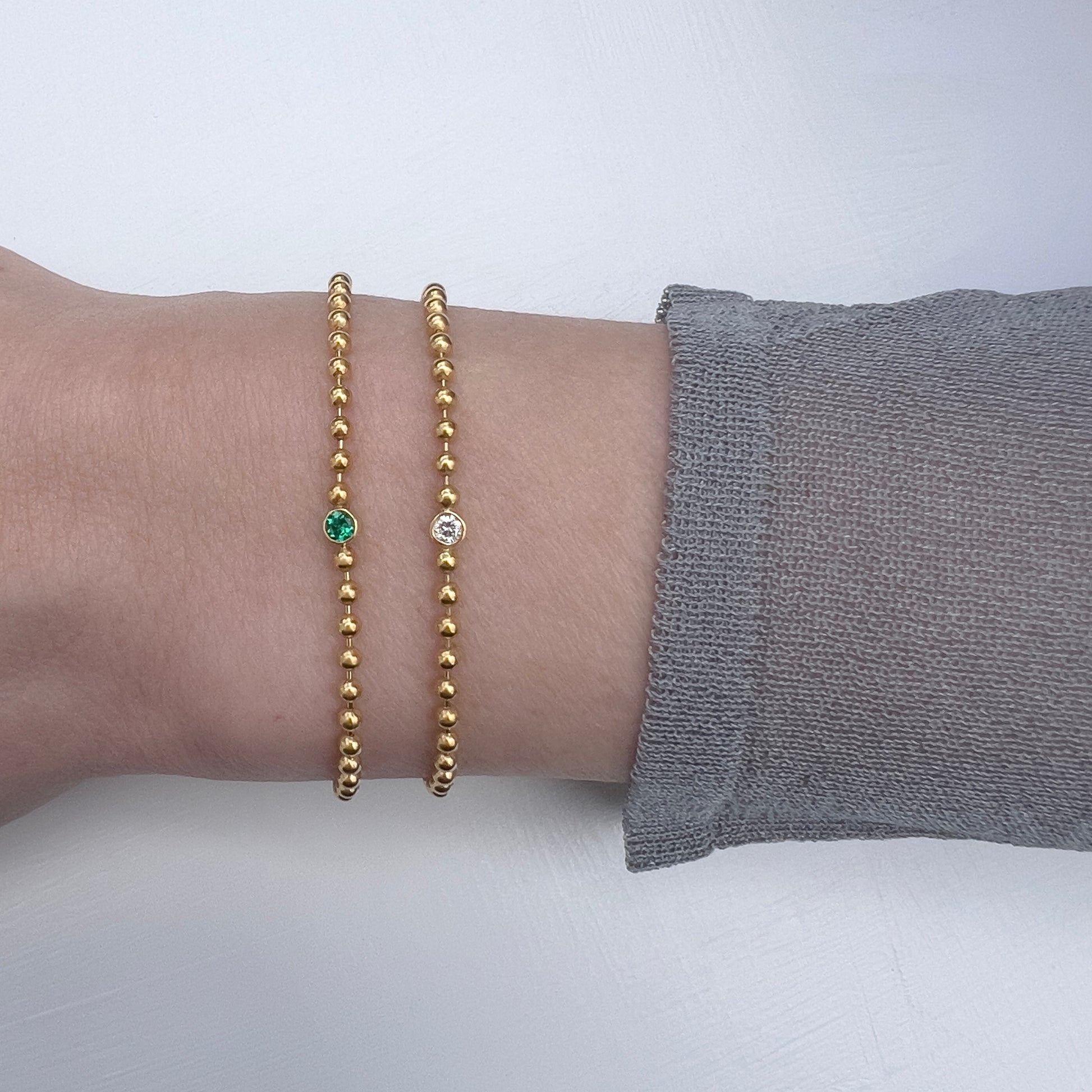 Arm wearing two gold ball chain bracelets set with emerald and diamonds.