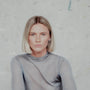 Person with blonde hair wearing silver hoop earrings and a gray sweater against a light gray background