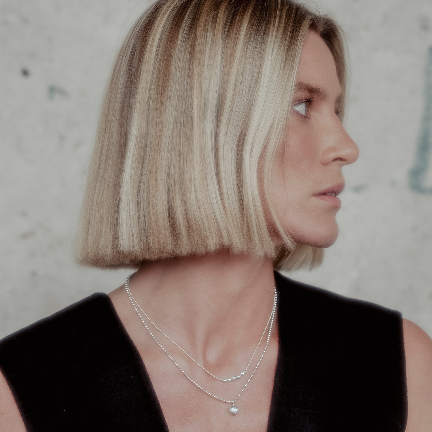 Woman with a bob haircut wearing a black top and layered silver orb necklaces against a neutral background