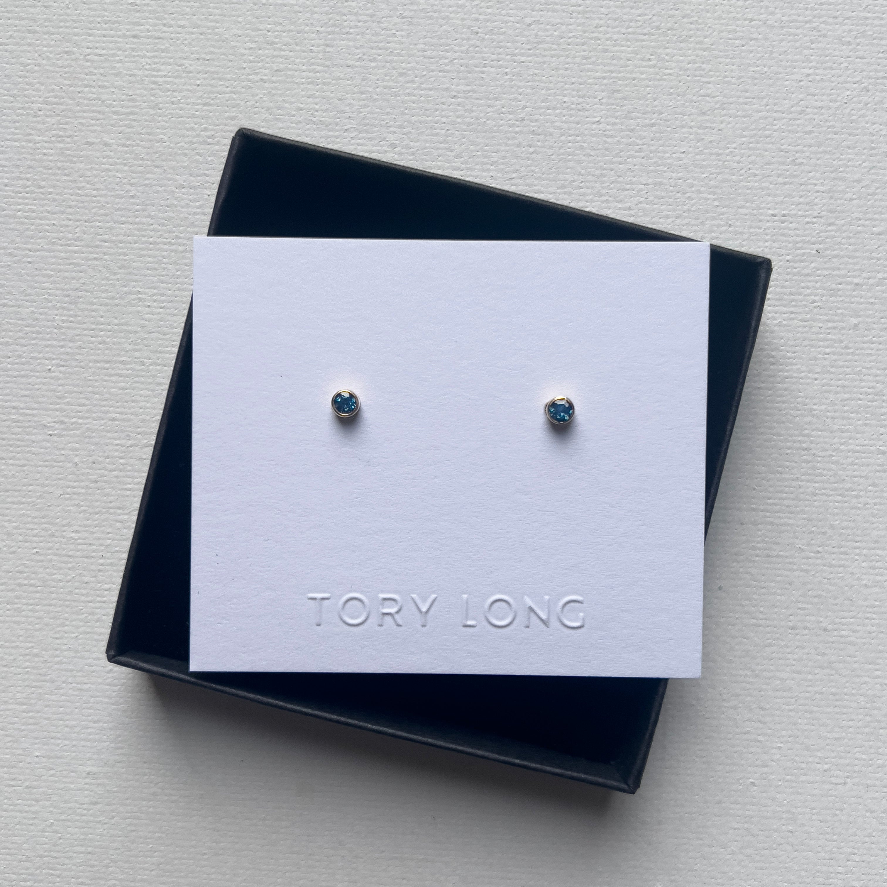 Pair of blue topaz birthstone earrings on a Tory Long branded card in a black box on a light gray background